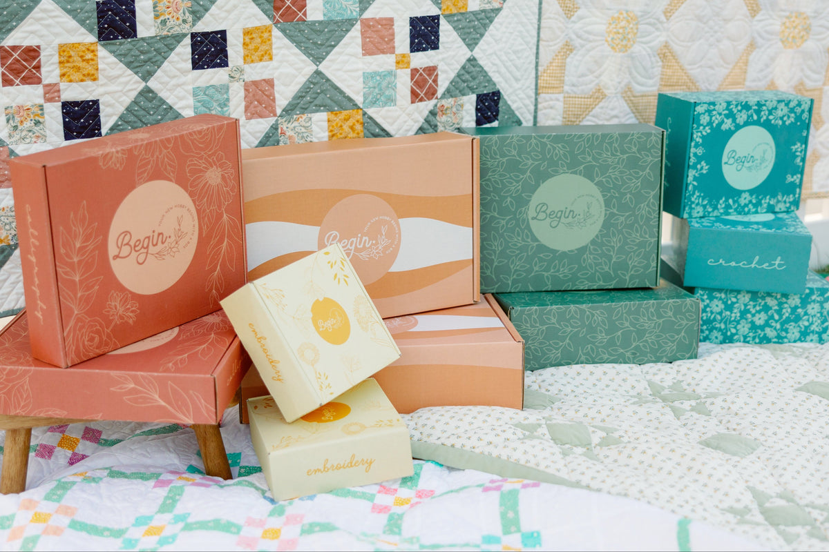 A collection of decorative product boxes from the brand Begio, displayed on a bed with a colorful quilted background. The boxes come in various pastel and earthy tones including coral, peach, sage green, and cream, with some featuring floral patterns and others displaying the circular Begio logo.